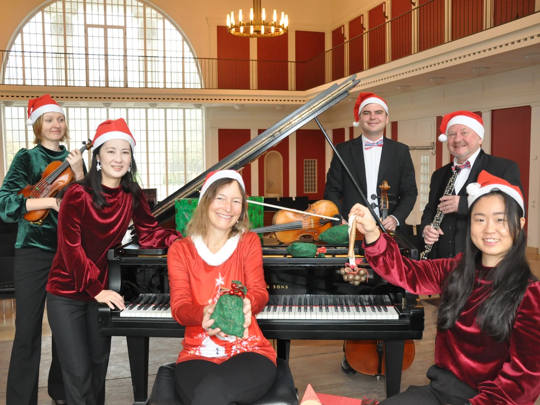 Adventskonzerte mit dem Staatsbad Orchester Musiker in roten Samt-Weihnachtsshirts und Mützen posieren mit Instrumenten vor einem Flügel in einem Saal.