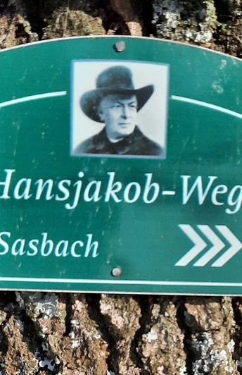 Grünes kleines Wanderschild Hansjakob-Weg am Baum befestigt, mit Wegweiser Richtung Sasbach.