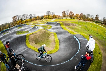 Ein geschwungener Pumptrack mit Radfahrern im herbstlichen Umfeld, umrahmt von Bäumen und Wiesen.