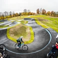 Ein geschwungener Pumptrack mit Radfahrern im herbstlichen Umfeld, umrahmt von Bäumen und Wiesen.