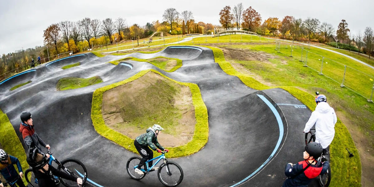 Ein geschwungener Pumptrack mit Radfahrern im herbstlichen Umfeld, umrahmt von Bäumen und Wiesen.