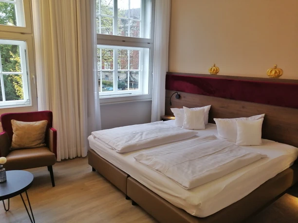 Helles Hotelzimmer mit zwei Einzelbetten, roten Akzenten und großen Fenstern mit Blick ins Grüne.