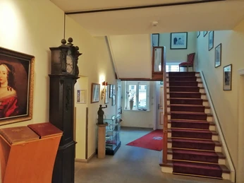 Ein Hotelinnenbereich mit altem Uhrschrank, Treppe mit rotem Teppich und Wandbildern.