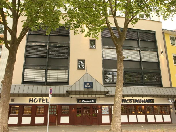 Hotel Münchner Hof-Herford (11)-Aussenansicht.JPG Dreistöckiges Gebäude mit heller Fassade und dunklen Fensterläden, beschattet von großen Bäumen.