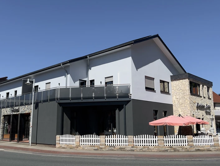 Ferienwohnungen "Orchideen Garten" Zweistöckiges weiß-graues Gebäude mit umlaufendem Balkon, rosa Sonnenschirm und weißem Zaun.