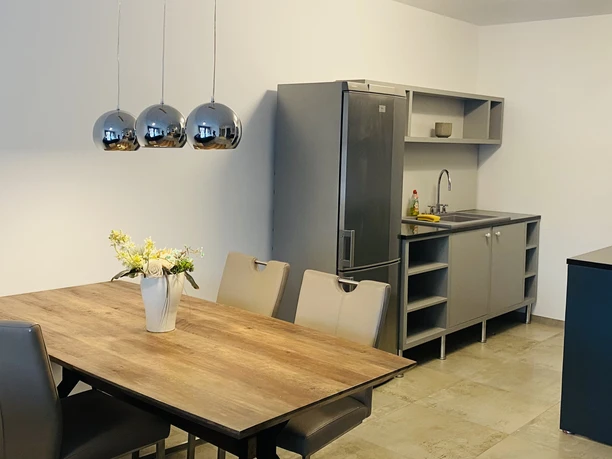 Ferienwohnungen "Orchideen Garten" - Küche/FeWo1 Moderner Küchen- und Essbereich mit Holztisch, vier Stühlen, Edelstahl-Kühlschrank und Hängelampen.