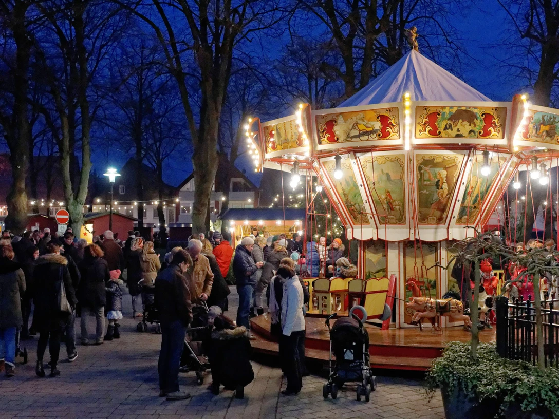 ._Weihnachtsmarkt_MWoestheinrich.jpg Ein nostalgisches Karussell dreht sich inmitten einer Menschenmenge auf einem stimmungsvollen Abendmarkt.
