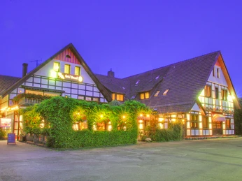 Hotel Vivendi-Herford-Aussenansicht.jpg Traditionelles Fachwerk-Hotel beleuchtet in der Abenddämmerung, umgeben von grünen Pflanzen.