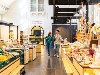 herford-markthalle-teutoburger-wald-tourismus-h-tornow-008-jpg.jpg Ein Paar schlendert durch die belebte Markthalle, umgeben von bunten Ständen und blühenden Blumen.