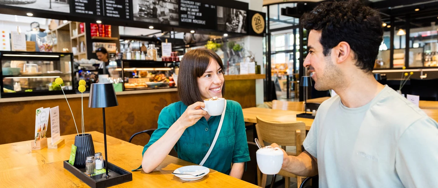 Ein Mann und eine Frau genießen Kaffee an einem hellen Holztisch in einer modernen Markthallenatmosphäre.