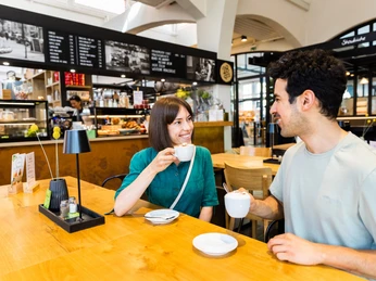herford-markthalle-teutoburger-wald-tourismus-h-tornow-022-jpg.jpg Ein Mann und eine Frau genießen Kaffee an einem hellen Holztisch in einer modernen Markthallenatmosphäre.