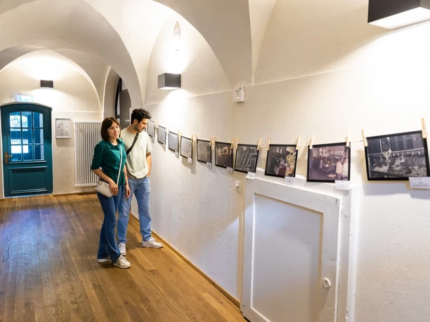 Personen betrachten eine Ausstellung historischer Fotos in einem langen, gewölbten Flur.