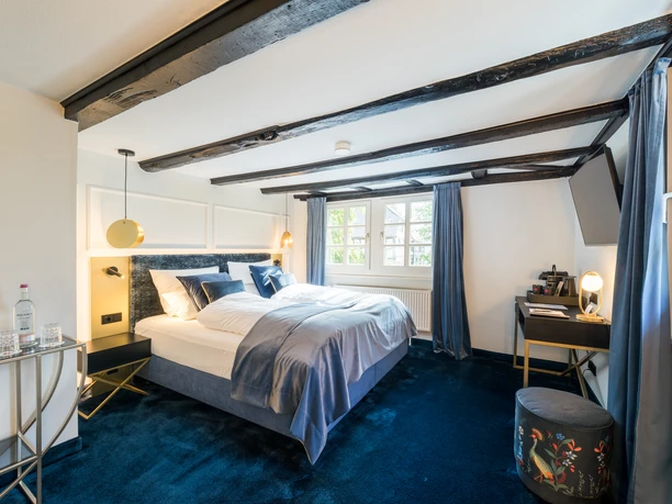 Hotel von Landsberg Gemütliches Schlafzimmer mit Doppelbett, blauen Akzenten, Holzdeckenbalken und zwei Fenstern mit Tageslicht.