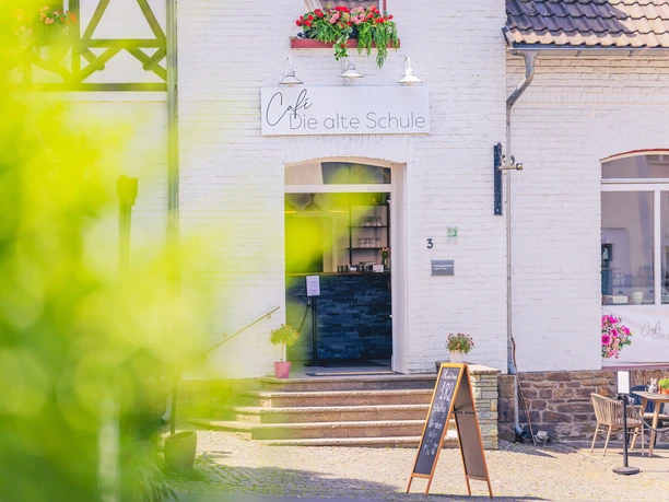 Café Die alte Schule Café "Die alte Schule" in einem charmanten weißen Gebäude mit blühenden Pflanzen und Holztür.