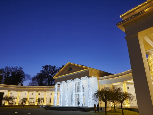 Beleuchtete Wandelhalle.jpg Hellerleuchtete Wandelhalle in der Abenddämmerung, mit festlicher Dekoration und Lichterketten.