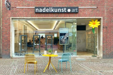 Fassade eines Geschäftes mit der Aufschrift „nadelkunst art“ und großem Schaufenster. Auf dem gepflasterten Gehweg davor stehen zwei Stühle an einem runden Tisch.