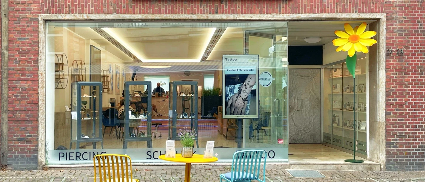 Nadelkunst.jpg Fassade eines Geschäftes mit der Aufschrift „nadelkunst art“ und großem Schaufenster. Auf dem gepflasterten Gehweg davor stehen zwei Stühle an einem runden Tisch.