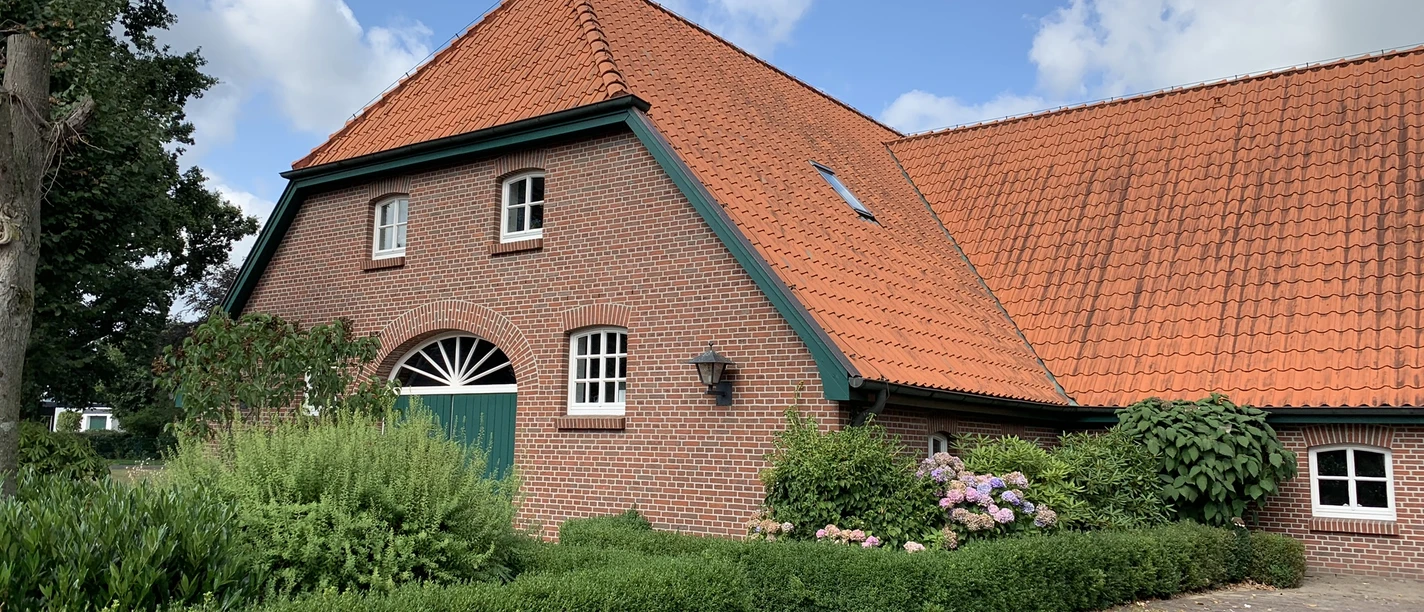 Hof Kleiberg Ein rotes Backsteinhaus mit spitzem Dach, umgeben von grünen Hecken und bunten Blumen, vor blauem Himmel.