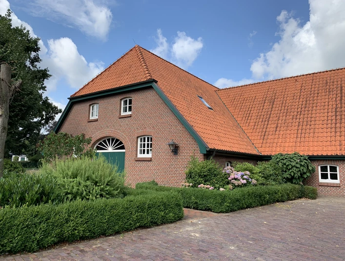 Hof Kleiberg Ein rotes Backsteinhaus mit spitzem Dach, umgeben von grünen Hecken und bunten Blumen, vor blauem Himmel.