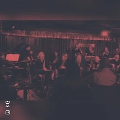 Eine Band spielt live in gedämpft rotem Licht vor einem sitzenden Publikum in einem Club.