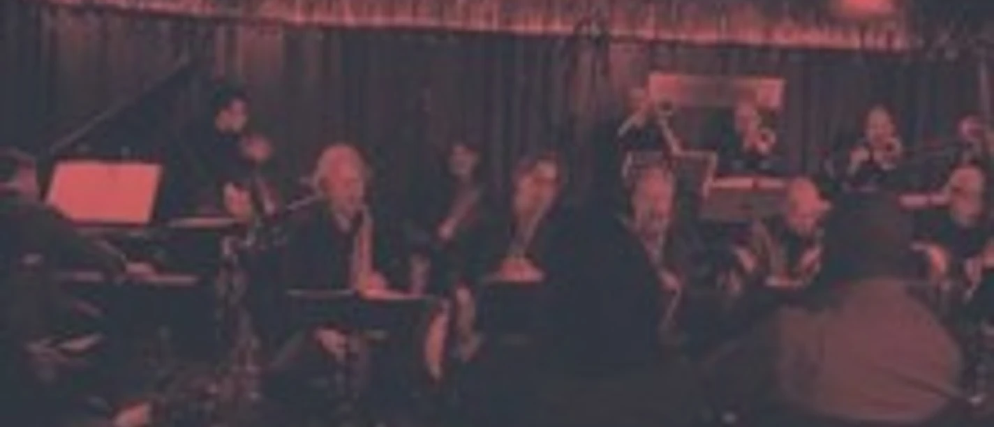 Eine Band spielt live in gedämpft rotem Licht vor einem sitzenden Publikum in einem Club.