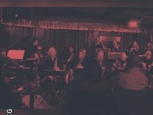 Eine Band spielt live in gedämpft rotem Licht vor einem sitzenden Publikum in einem Club.