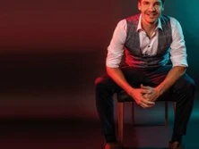 Ein lächelnder Mann sitzt in einem stilvollen Studio, trägt ein weißes Hemd mit grauer Weste.