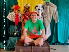 Ein fröhlicher Mensch in buntem Outfit sitzt vor einer Kulisse mit lustigen, tierischen Kostümen.