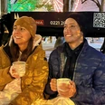 Rikolonia Köln Wintertouren Glühwein Rikscha.png Ein Pärchen sitzt lächelnd in einer Rikscha und jeder hält eine weiße Tasse in der HandA couple sits smiling in a rickshaw, each holding a white cup