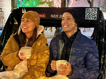 Rikolonia Köln Wintertouren Glühwein Rikscha.png Ein Pärchen sitzt lächelnd in einer Rikscha und jeder hält eine weiße Tasse in der HandA couple sits smiling in a rickshaw, each holding a white cup