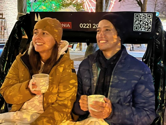 Rikolonia Köln Wintertouren Glühwein Rikscha.png Ein Pärchen sitzt lächelnd in einer Rikscha und jeder hält eine weiße Tasse in der HandA couple sits smiling in a rickshaw, each holding a white cup