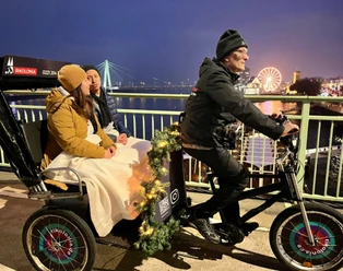 Rikolonia Köln Wintertouren II.png Rikscha fährt über eine Brücke über den Rhein und transportiert zwei Personen