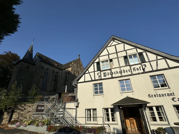 Dürscheider Hof - Außenansicht Restaurant & Kirche <p>Fachwerkhaus "Büscheder Hof" neben einer Kirche in Kürten, unter blauem Himmel, umgeben von Blumen.</p>