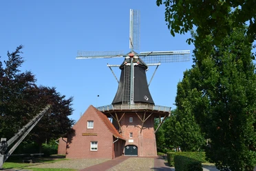 Kreutzmanns Mühle Werlte quer.JPG Historische Windmühle mit Backsteingebäude unter blauem Himmel, umgeben von grünen Bäumen.