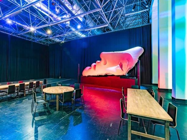 Oper Köln im StaatenHaus Das Bild zeigt einen modernen Probenraum einer Oper mit hohen Decken. Zentral platziert ist ein großes Kunstwerk in Form eines rosafarbenen Gesichtes, das auf einem Podest liegt. Umgeben ist der Raum von minimalistischen Holztischen und Stühlen, die im Kontrast zu den dramatischen Farben des Kunstwerks stehen.