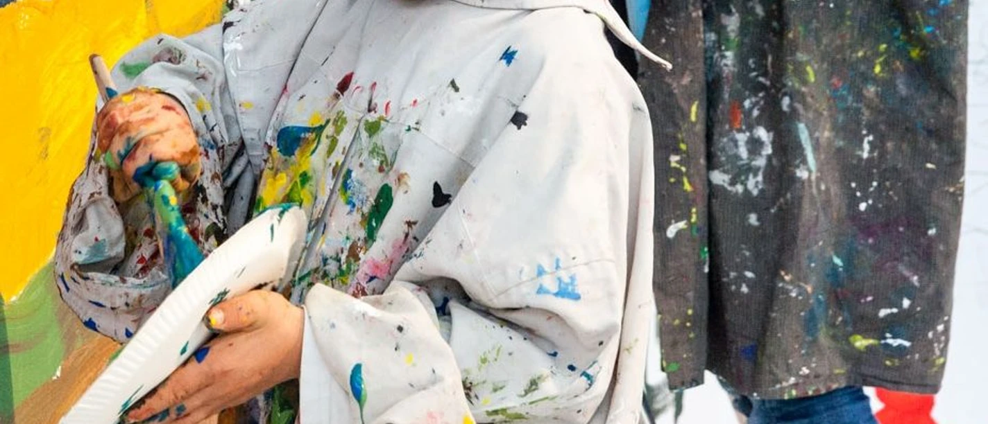 Sonntagsmalerei_LudwigForum-Foto-Carl-Brunn.jpg Ein Kind malt konzentriert an einer Leinwand, bekleidet mit einem übergroßen, buntspritzigen Malkittel.