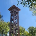 Holzturm mit Aussichtsplattform im Grünen; helle Sonne und umgebende Laubkronen rahmen die Ansicht.
