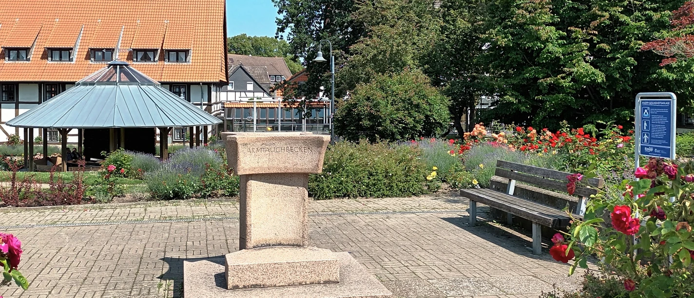 Steinernes Armtauchbecken auf einem gepflasterten Platz im Rosengarten vor einem Fachwerkhaus.
