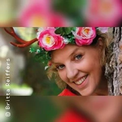 Lächelnde Frau mit Blütenkranz und kleinen Geweih-Elementen lehnt an einem Baum.