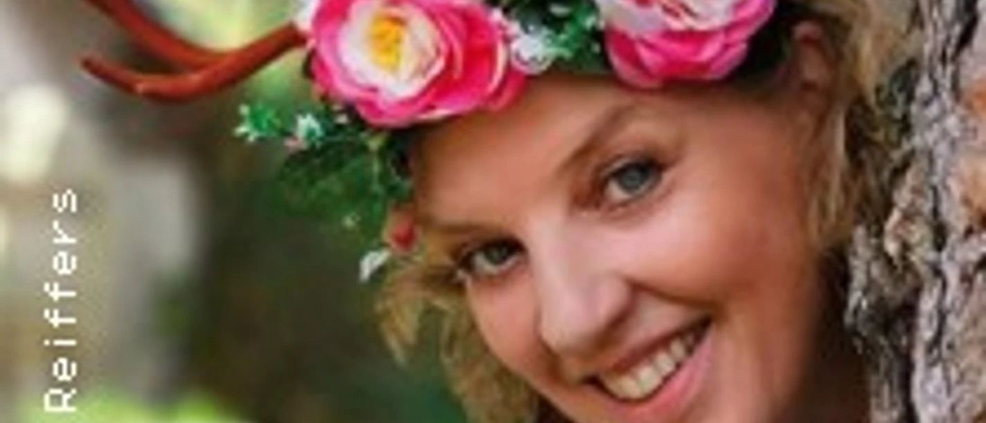 Lächelnde Frau mit Blütenkranz und kleinen Geweih-Elementen lehnt an einem Baum.
