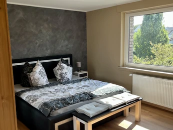 Schlafzimmer der Ferienwohnung im Weserbergland Modernes Schlafzimmer mit schwarzem Doppelbett, stilvoller Bettwäsche in schwarz-weiß Musterung, grauer Wischtechnik-Tapete und vor dem Bett eine Sitzbank aus Holz.