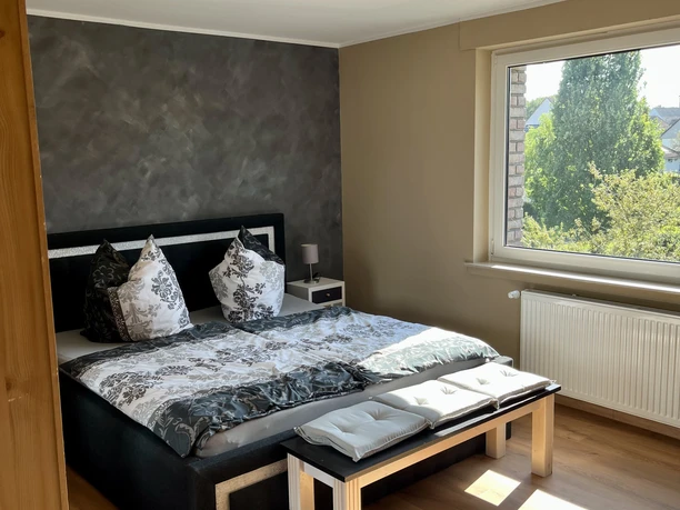 Schlafzimmer der Ferienwohnung im Weserbergland Modernes Schlafzimmer mit schwarzem Doppelbett, stilvoller Bettwäsche in schwarz-weiß Musterung, grauer Wischtechnik-Tapete und vor dem Bett eine Sitzbank aus Holz.