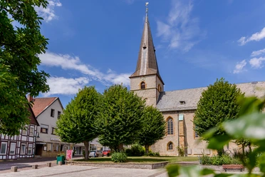 Ev.-Luth. St. Jacobi Kirche Werther Kirche mit hohem Turm umgeben von Bäumen, historisches Fachwerkhaus rechts, blauer Himmel.