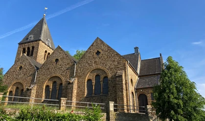 Braune Backsteinkirche mit hohen Giebeln und Kirchturm vor blauem Himmel, umgeben von Bäumen.
