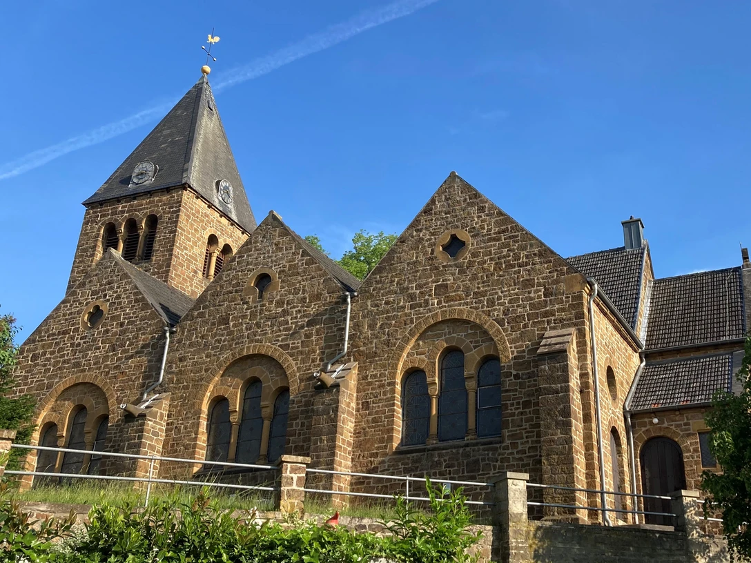 ev. Kirche Bad Holzhausen Braune Backsteinkirche mit hohen Giebeln und Kirchturm vor blauem Himmel, umgeben von Bäumen.