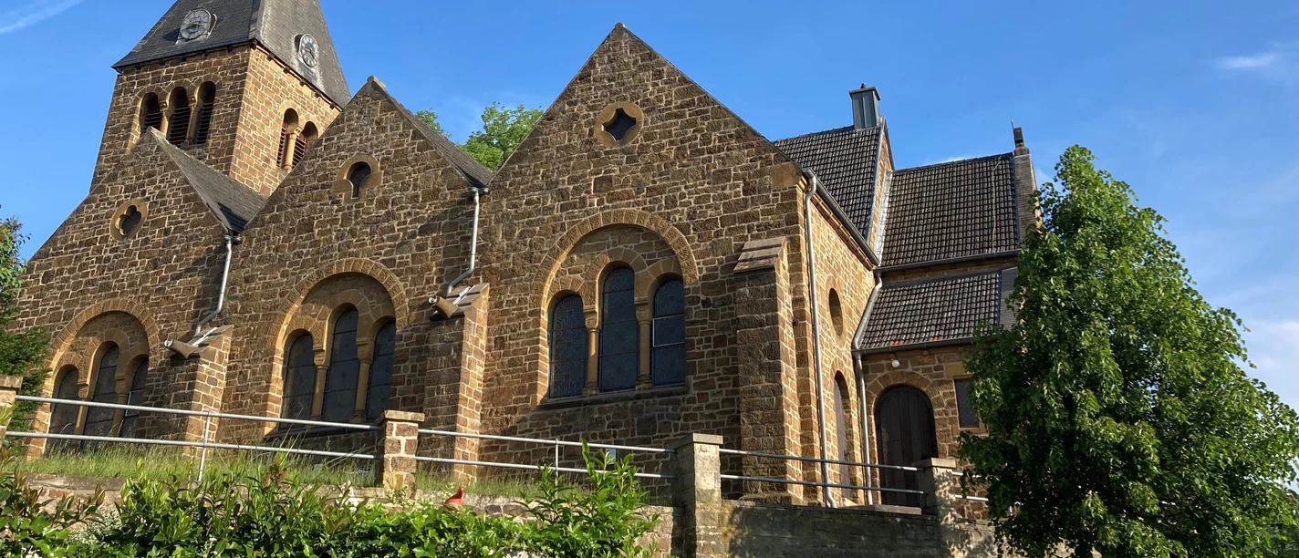 ev. Kirche Bad Holzhausen Braune Backsteinkirche mit hohen Giebeln und Kirchturm vor blauem Himmel, umgeben von Bäumen.
