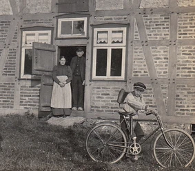 geschichte_auf_raedern-900009941-22700-11.webp Ein Junge mit Fahrrad vor einem Fachwerkhaus; im Hintergrund schauen zwei Erwachsene aus der Tür.