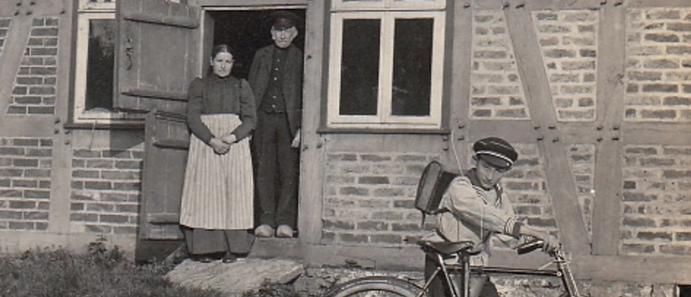 geschichte_auf_raedern-900009941-22700-11.webp Ein Junge mit Fahrrad vor einem Fachwerkhaus; im Hintergrund schauen zwei Erwachsene aus der Tür.