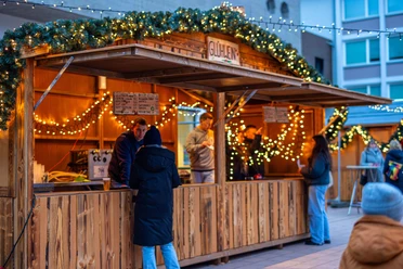 Sülzer Weihnachtsdorf Ein beleuchteter Weihnachtsmarktstand mit Personen und dekorativen Lichtern.