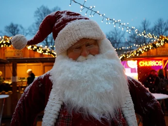 Sülzer Christmas Village Weihnachtsmann mit weißem Bart und roter Mütze, umgeben von festlich beleuchteten Ständen.Santa Claus with a white beard and red hat, surrounded by festively illuminated stalls.